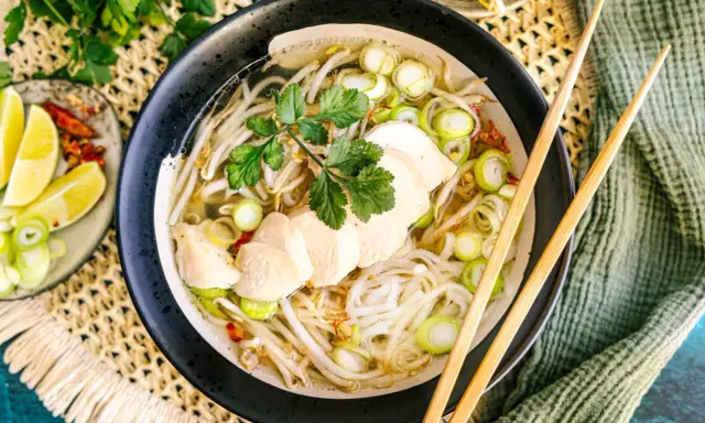 Eine dampfende Schüssel Pho Ga mit zarten Hühnerbrustscheiben, Reisnudeln, Sojasprossen und Frühlingszwiebeln. Serviert mit Limettenspalten und Chili.