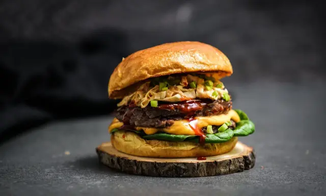 Ein saftiger koreanischer Burger mit doppeltem Patty, Käse, knackigem Krautsalat und Frühlingszwiebeln auf einem rustikalen Holzbrett.