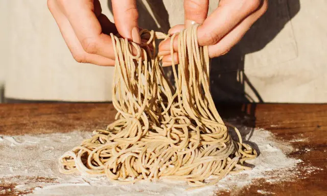 Hände halten frisch gemachte Ramen-Nudeln über einem Holztisch mit Mehl. So einfach kann ramen selber machen sein!