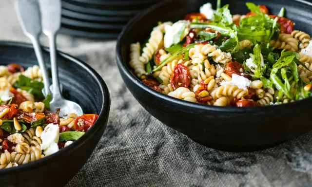 Zwei Schalen mit einem gesunden Nudelsalat aus Spiralnudeln, getrockneten Tomaten, Pinienkernen, Rucola und Feta.