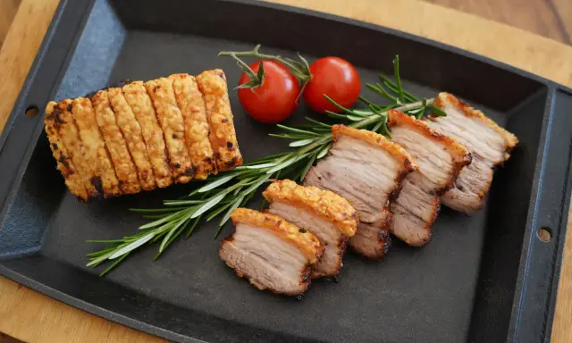 Knuspriger asiatischer Schweinebauch, in Scheiben geschnitten, mit Rosmarin und Kirschtomaten auf einer Gusseisenplatte serviert.