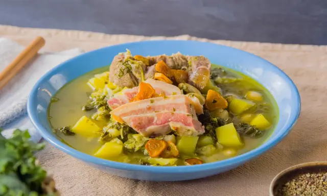 Herzhafter spanischer Eintopf mit Fleisch, Kartoffeln, Grünkohl und Karotten in einer blauen Schüssel.