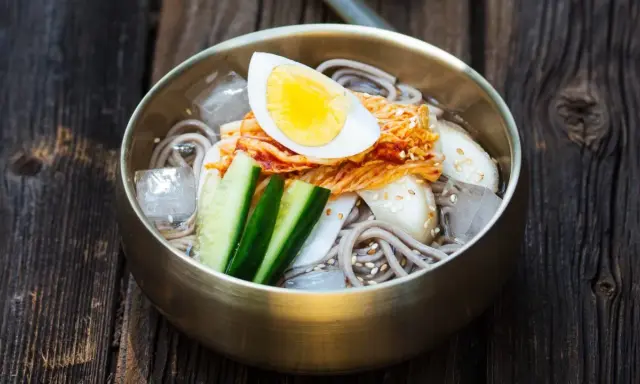 Erfrischendes Mul Naengmyeon mit Buchweizennudeln, Kimchi, Gurke, Ei und Eiswürfeln in einer bronzenen Schüssel auf Holzuntergrund.
