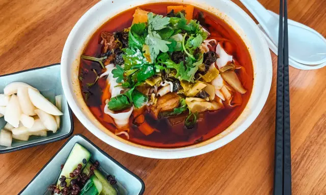 Eine Schüssel mit aromatischer taiwanischer Rindernudelsuppe, garniert mit frischem Koriander, daneben eingelegte Gurken und Rettich.