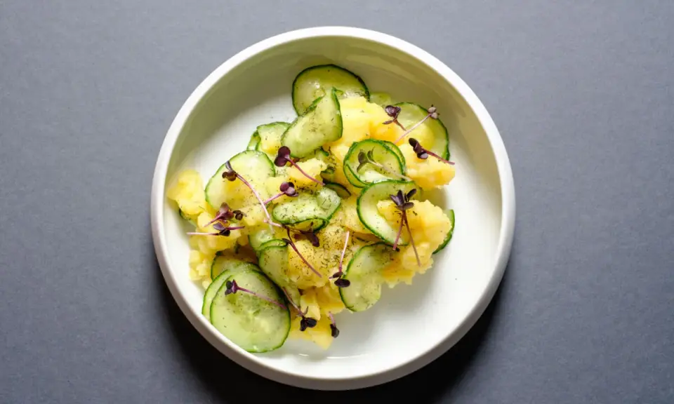 Kartoffelsalat mit frischer Gurke, garniert mit lila Sprossen und Dill. Ein leichter und erfrischender Genuss.