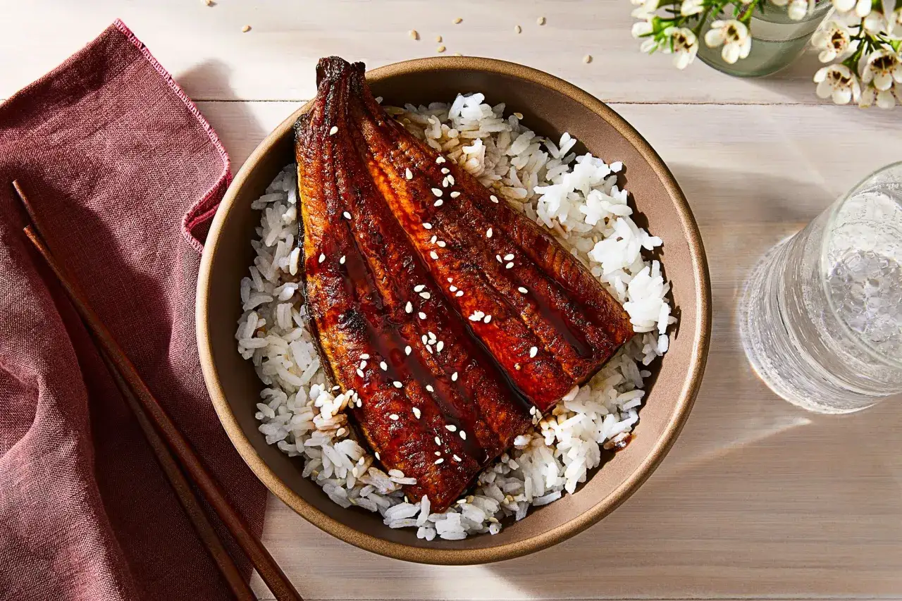 Schmackhaftes Unagi (Aal) mit süßer Soße auf Reis, bestreut mit Sesam. Ein Glas Wasser und Stäbchen liegen daneben.