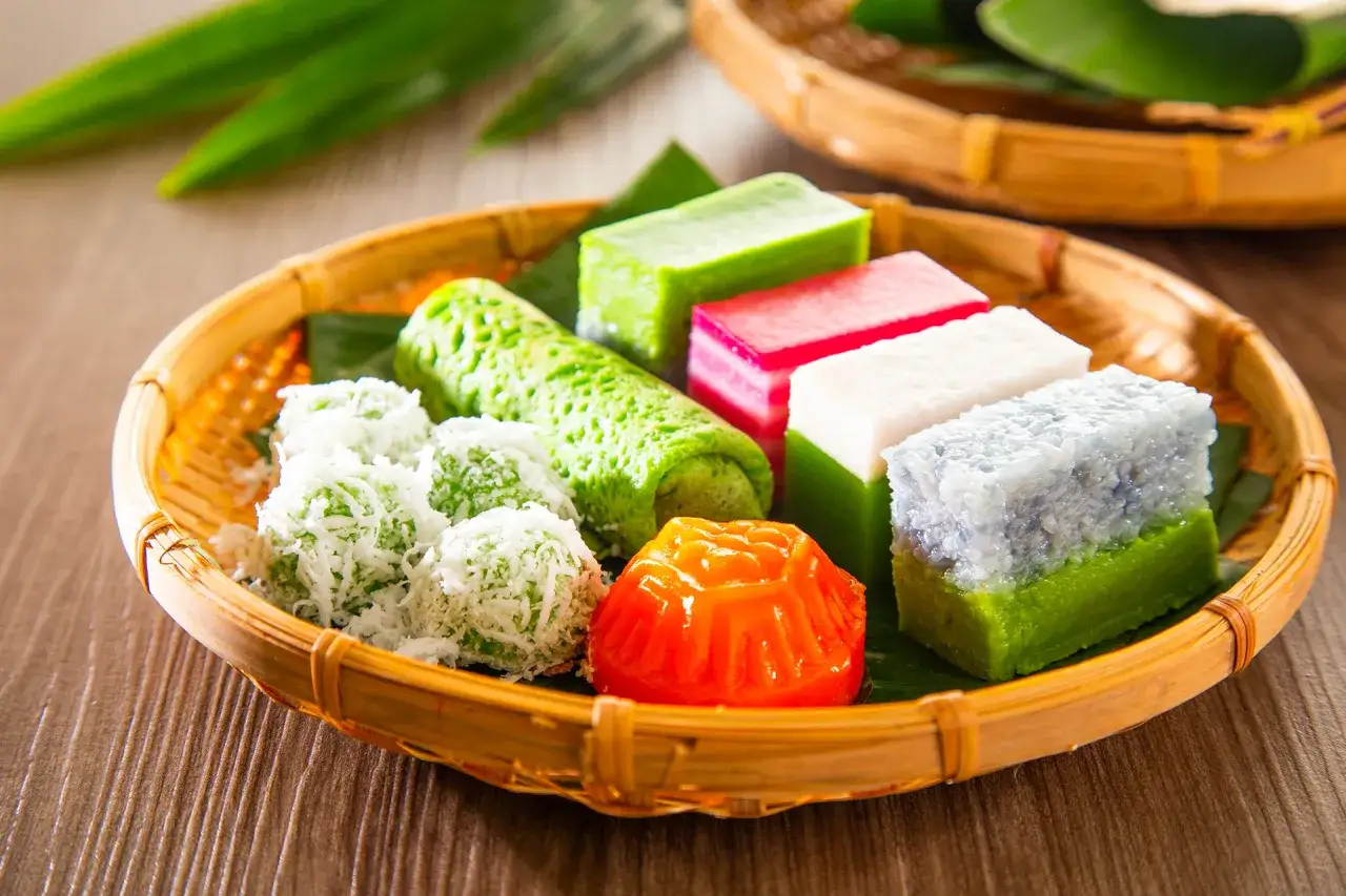 Bunte asiatische Nachspeisen wie grüne Röllchen, Kokoskugeln und Schichtkuchen in einem Korbe.
