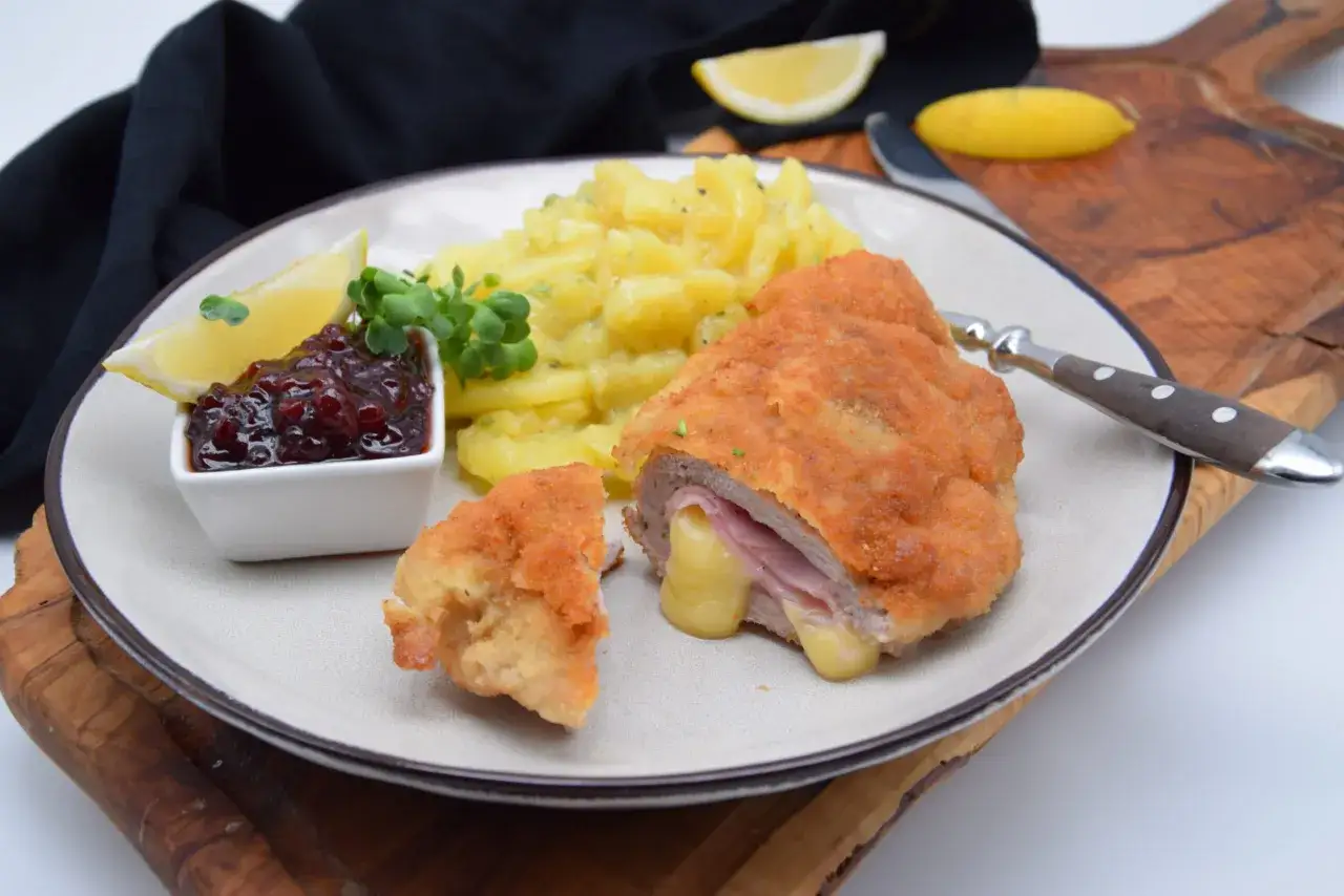 Ein knuspriges Cordon Bleu mit geschmolzenem Käse, dazu Kartoffelsalat, Preiselbeeren und Zitrone auf einem Teller.
