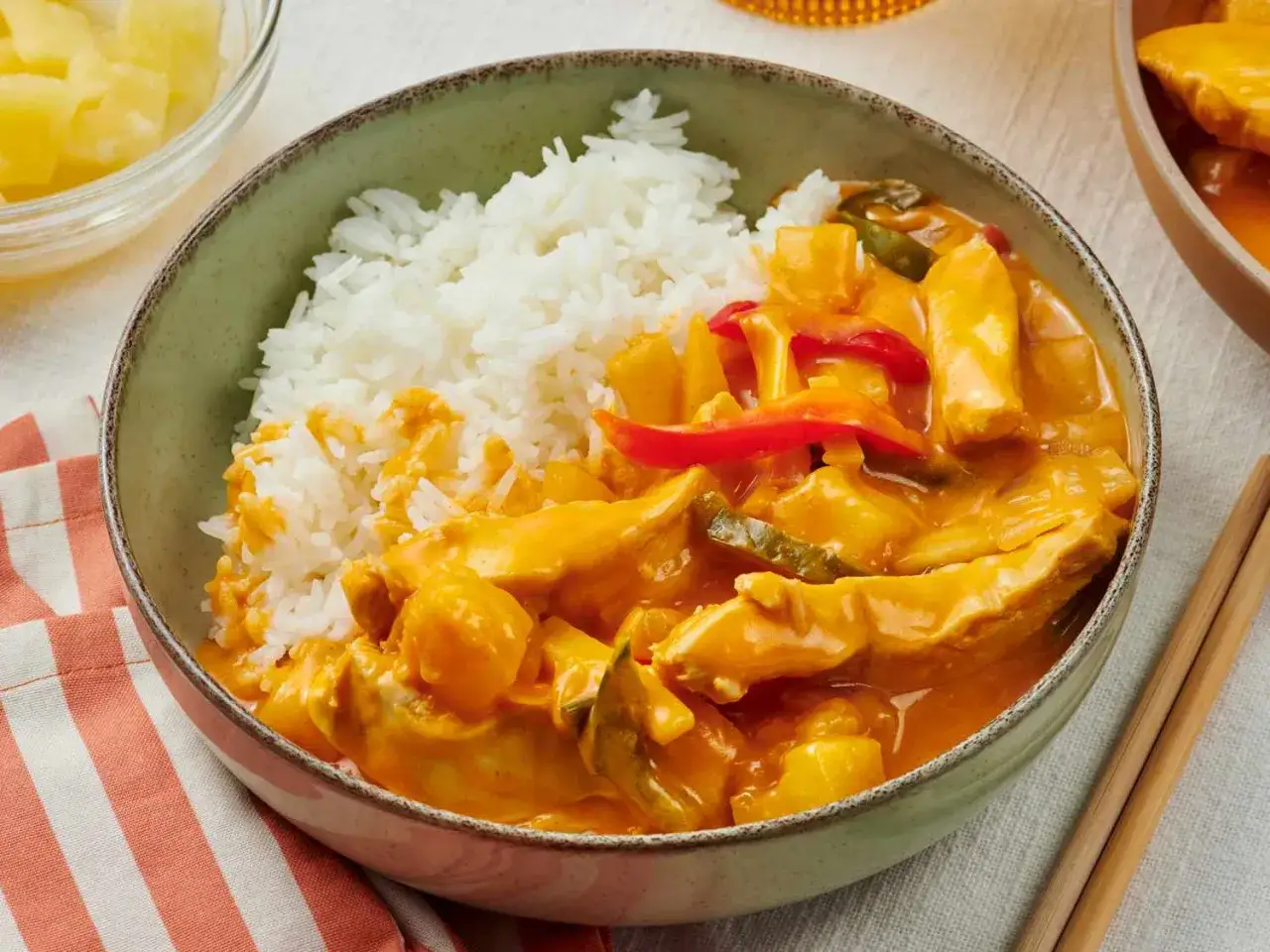 Schüssel mit Reis und cremigem thai curry mit ananas und kokosmilch, garniert mit Paprika und Zucchini. Daneben ein Schälchen mit Ananasstücken.