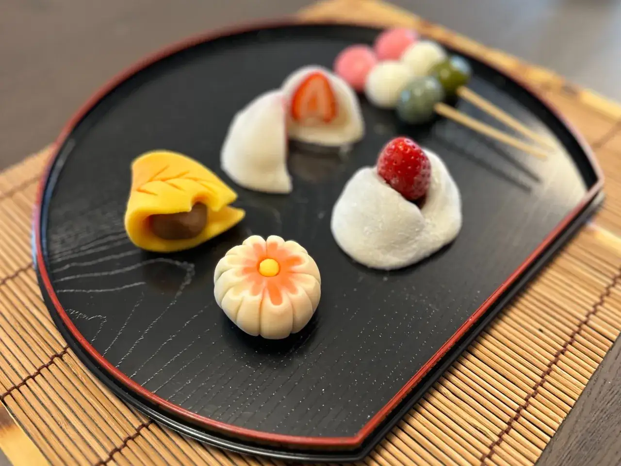 Bunte Wagashi-Süßigkeiten, inspiriert von einem Wagashi-Rezept, auf einem schwarzen Teller: Mochi mit Erdbeeren, ein Blatt mit Füllung und eine Blumenform.
