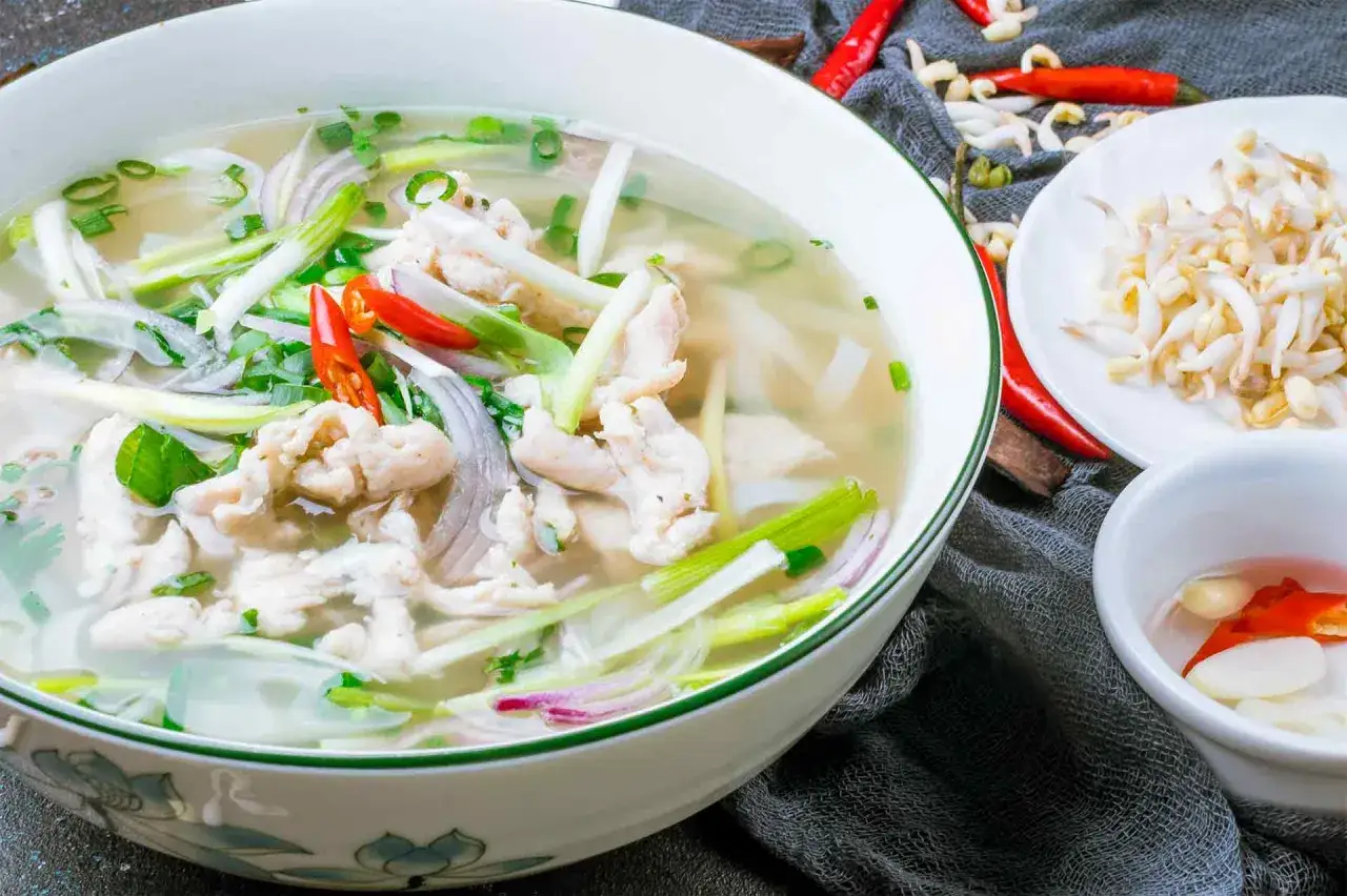 Eine Sch&uuml;ssel mit Canh Chua, einer vietnamesischen H&uuml;hnersuppe, garniert mit Fr&uuml;hlingszwiebeln und Chili. Daneben liegen Sprossen und eine kleine Schale mit Knoblauch und Chili.