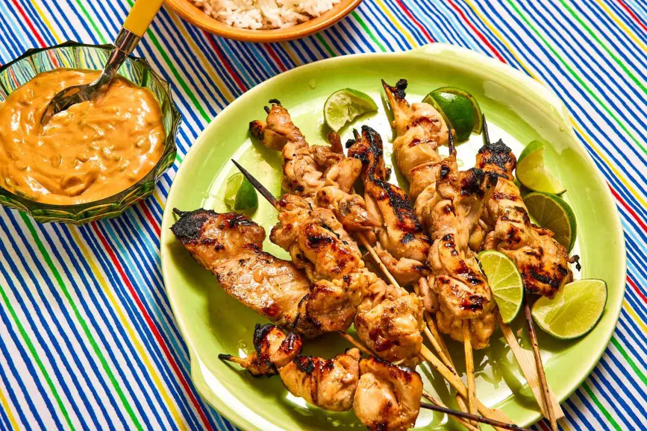 Saftige H&auml;hnchen-Satay-Spie&szlig;e mit Erdnusssauce und Limettenspalten auf einem bunten Tischl&auml;ufer.