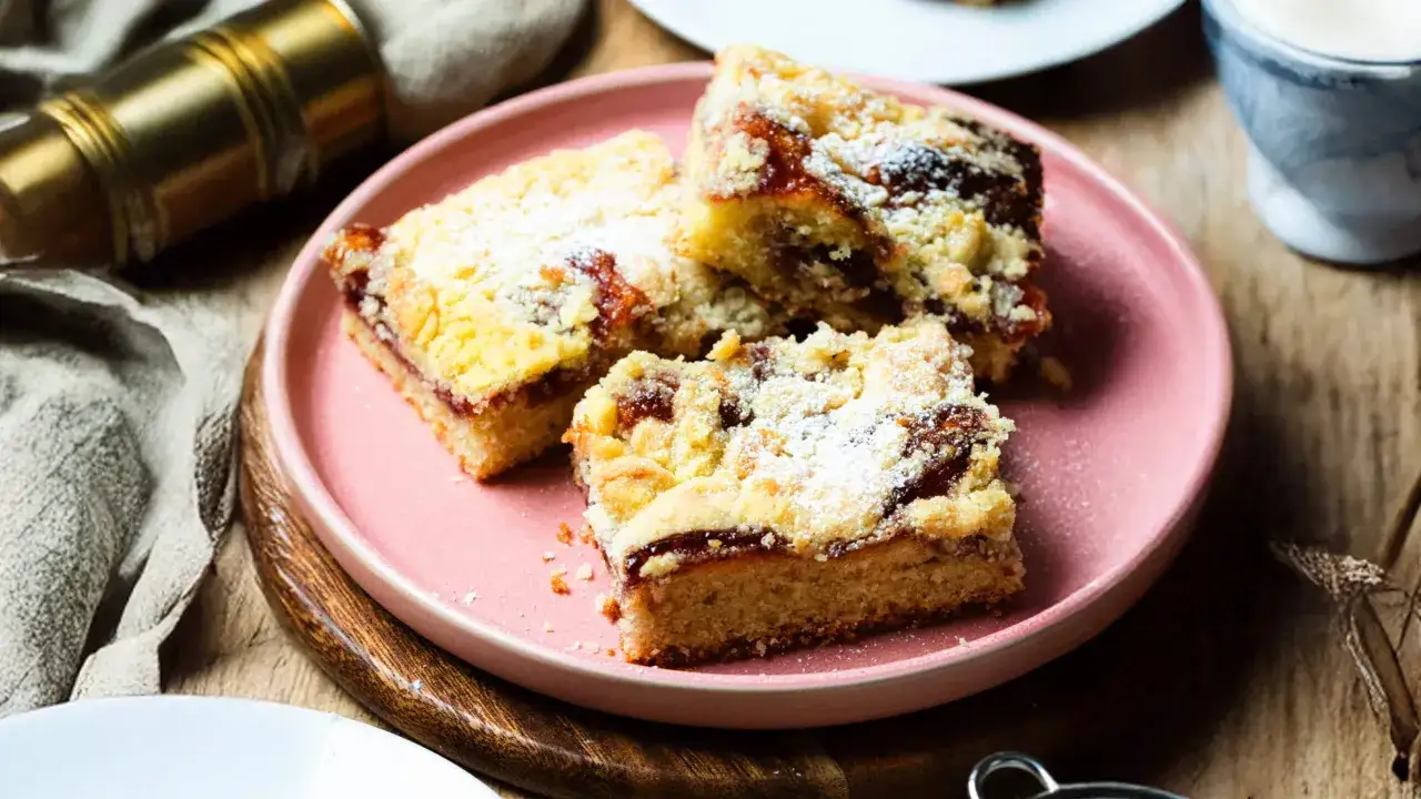 Saftiger Streuselkuchen mit Marmelade, best&auml;ubt mit Puderzucker, auf einem rosa Teller.