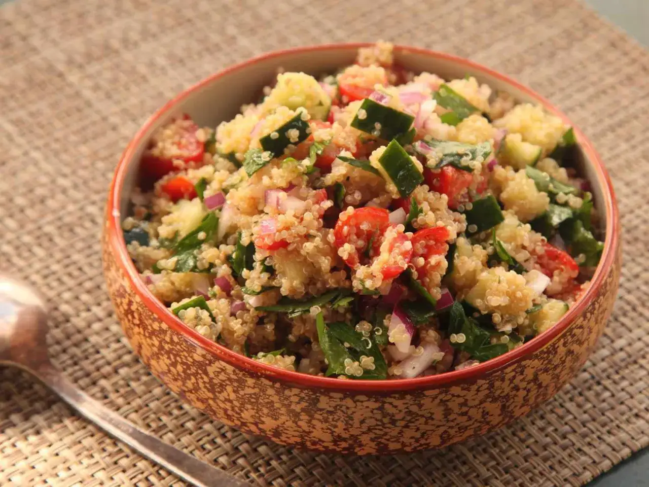 Schüssel mit Quinoa-Salat mit Feta, Tomaten, Gurken und roten Zwiebeln, serviert auf einer braunen Matte.