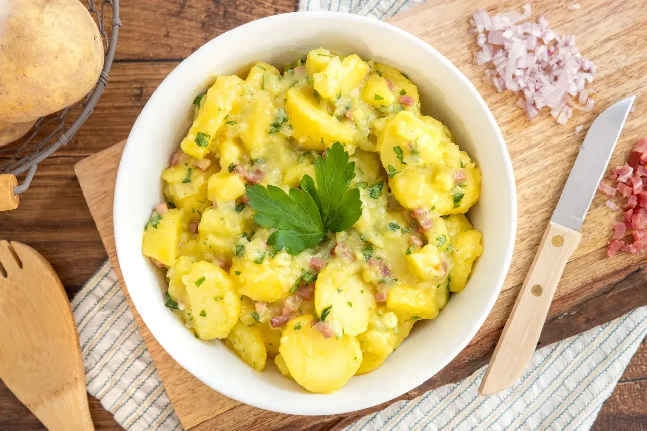 Ein klassisches Kartoffelsalat Rezept, appetitlich in einer Schüssel serviert, mit frischer Petersilie garniert. Daneben liegen Zutaten wie Speckwürfel und Zwiebeln.