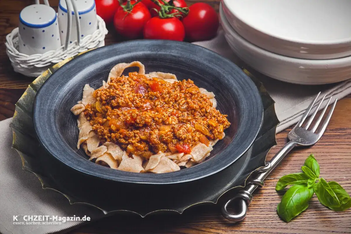 Ein Teller mit breiten Bandnudeln und einem herzhaften, italienischen Ragout. Frische Tomaten und Basilikum deuten auf die aromatische Zubereitung hin.