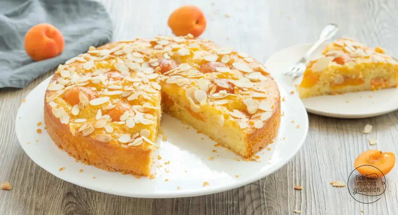 Saftiger aprikosenkuchen mit mandeln, garniert mit frischen Aprikosen und Mandelblättchen. Ein Stück ist bereits auf einem Teller angerichtet.