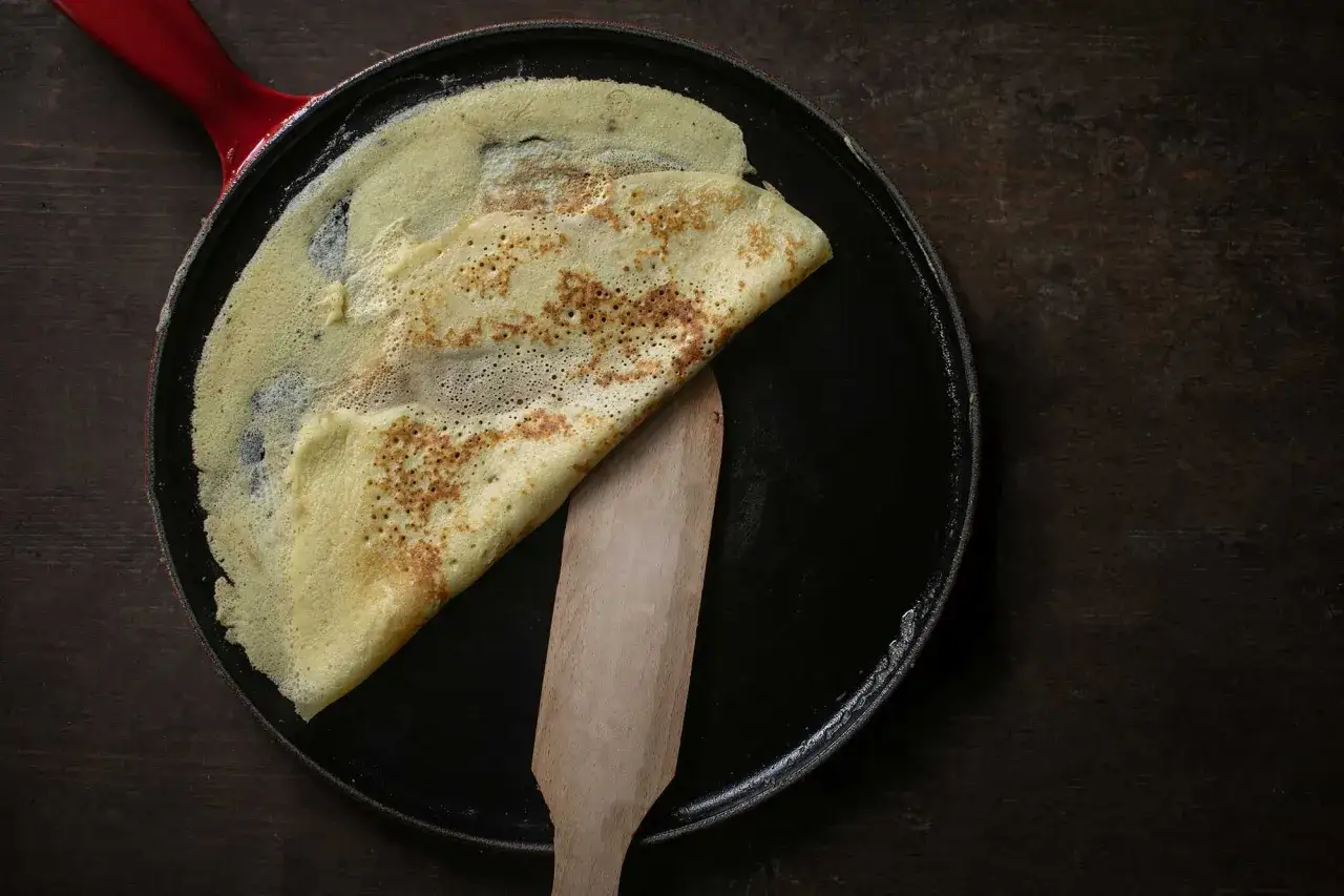 Ein goldbrauner Crêpe wird in einer Pfanne mit Holzspatel gewendet. Das perfekte Crepes Rezept original.