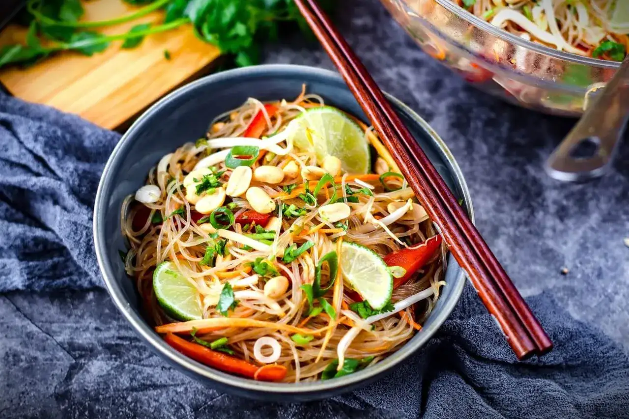 Schüssel mit Glasnudelsalat, garniert mit Erdnüssen, Limettenspalten und Koriander. Chopsticks liegen auf der Schüssel.