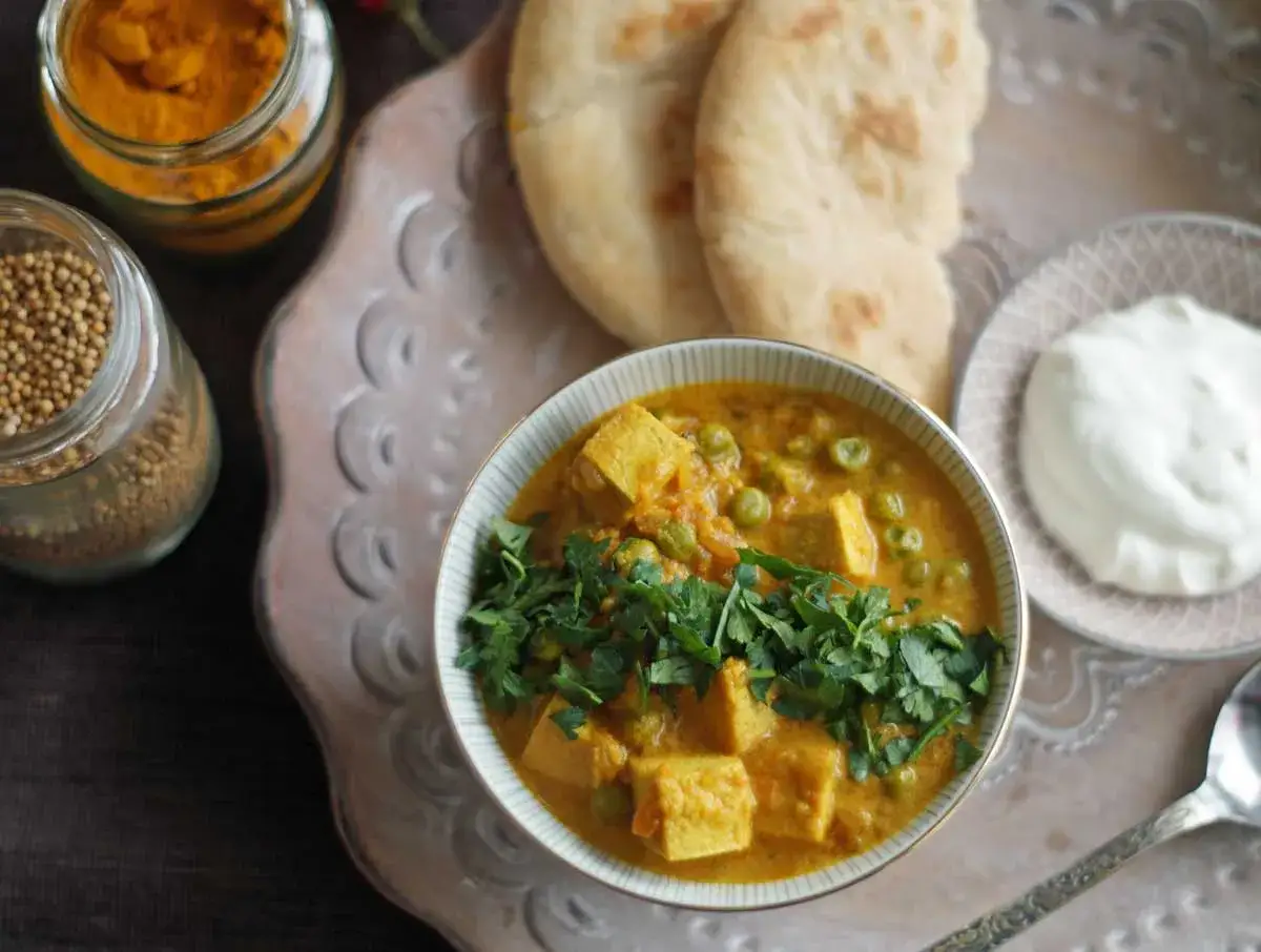 Schüssel mit cremigem Mattar Paneer, garniert mit Koriander. Daneben Naan, Joghurt und Gewürze.