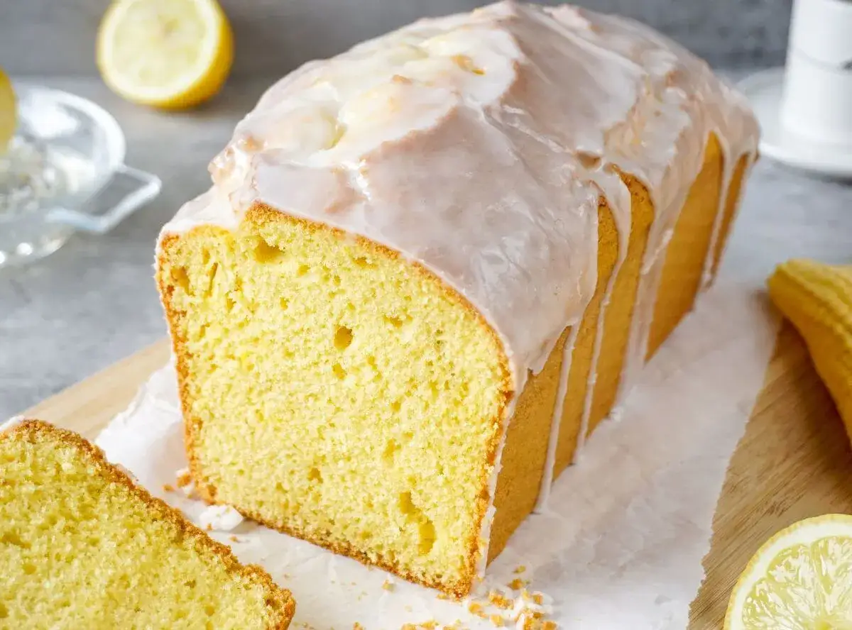 Ein fluffiger, saftiger Zitronenkuchen mit Zuckerguss, der auf einem Holzbrett liegt. Daneben liegen Zitronenscheiben.
