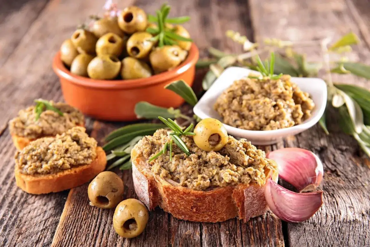 Herzhaftes Tapenade deutsch auf knusprigem Brot, garniert mit Oliven und Rosmarin. Daneben frischer Knoblauch und eine Schale Oliven.