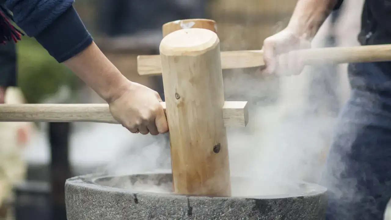 Hände schlagen mit Holzstößeln auf gedämpften Reis in einem Steinmörser. Ein traditionelles mochi rezept wird hier lebendig.