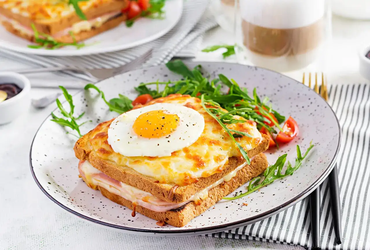 Ein köstlicher Croque Madame mit geschmolzenem Käse, Schinken und einem Spiegelei, serviert mit Rucola und Kirschtomaten.