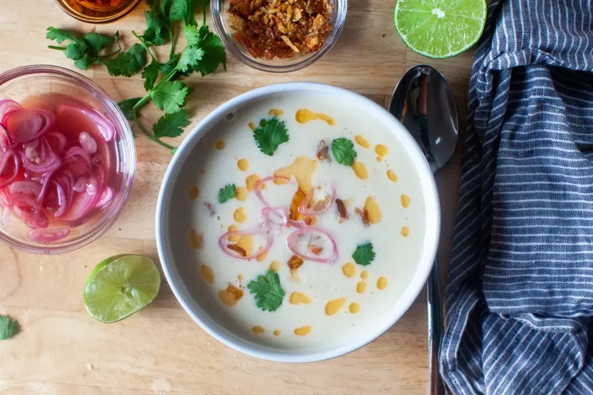 Cremige Maissuppe mit Kokosmilch, garniert mit roten Zwiebeln, Koriander und einem Hauch Chili&ouml;l. Dazu gibt es Limettenspalten und knusprige R&ouml;stzwiebeln.