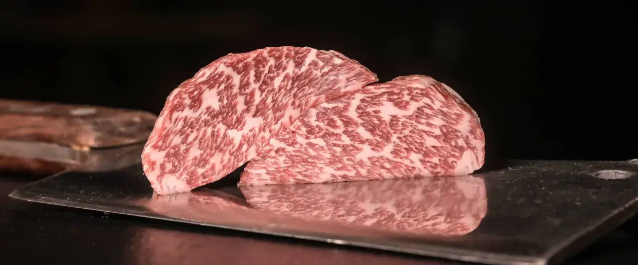 Zwei St&uuml;cke Kobe Rindfleisch mit charakteristischem Marmormuster liegen auf einem Metallmesser. Das Aussehen des Kobe Rindfleischs ist unverwechselbar.
