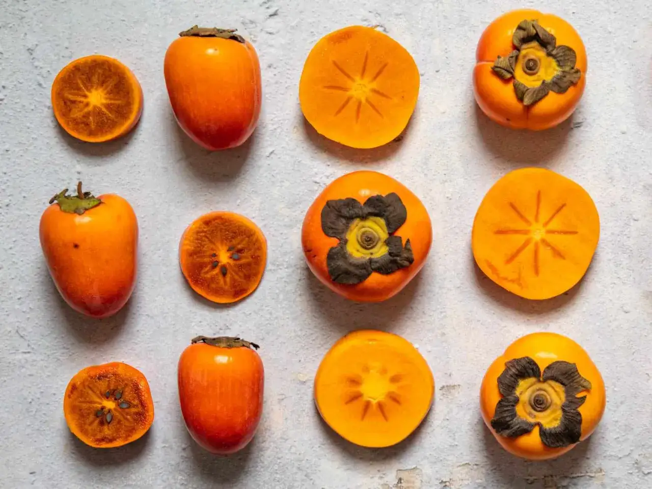 Ganze und halbierte Kakis liegen auf hellem Untergrund. Sie sind leuchtend orange und zeigen ihre Kerne. Sind Kakis gesund? Ja, sie sind voller Vitamine!
