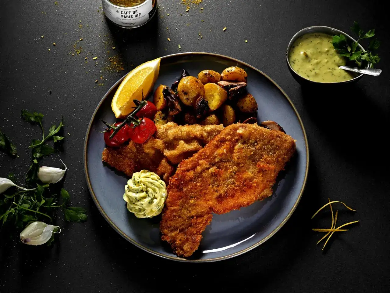 Ein knuspriges Wiener Schnitzel mit Bratkartoffeln, Tomaten, Zitrone und Kräuterbutter. Dazu eine cremige Sauce.