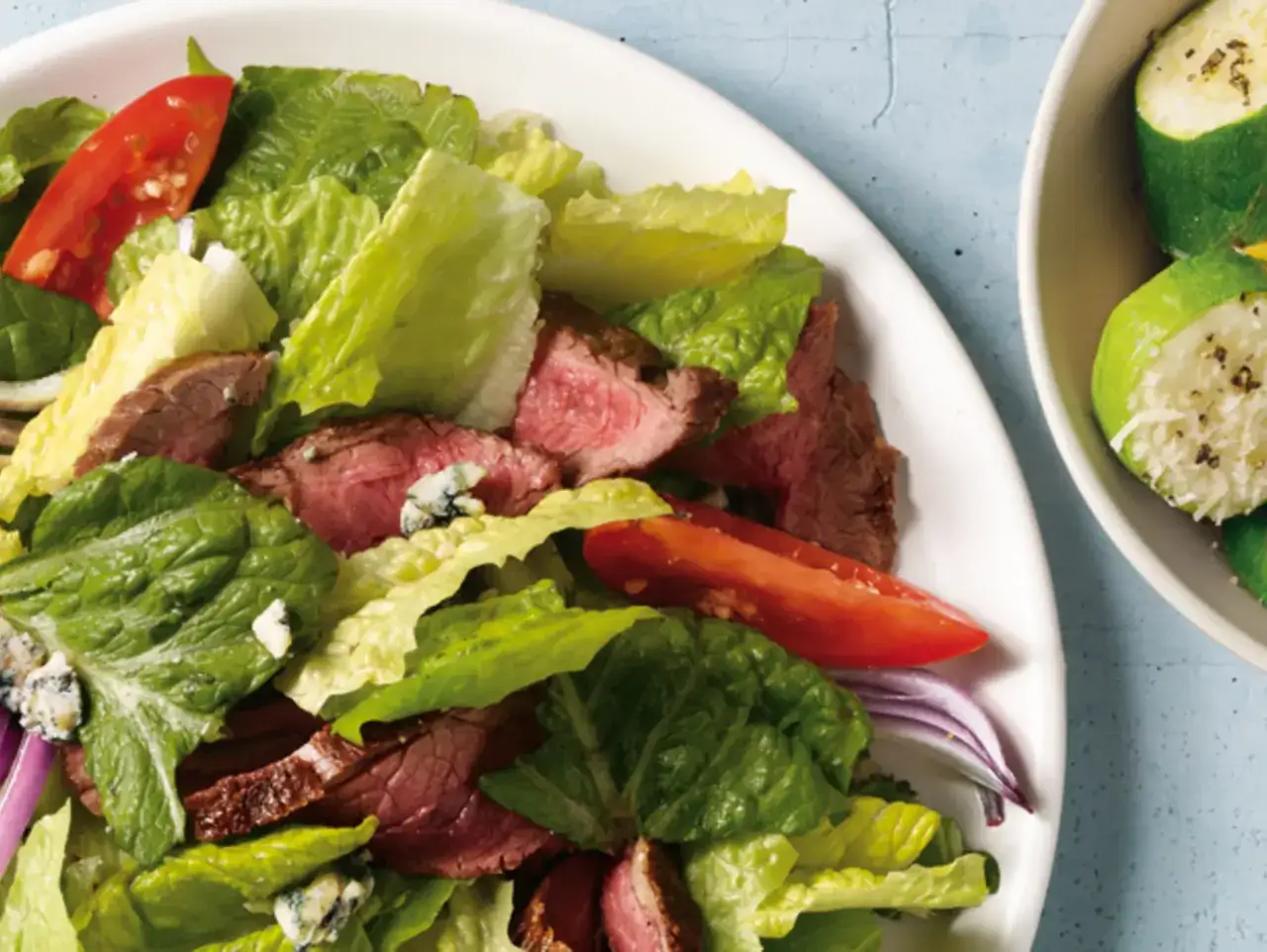 Saftiger Steak Salat mit knackigem Salat, Tomaten, Zwiebeln und Blauschimmelk&auml;se. Daneben ein Sch&auml;lchen mit gegrilltem Zucchini.