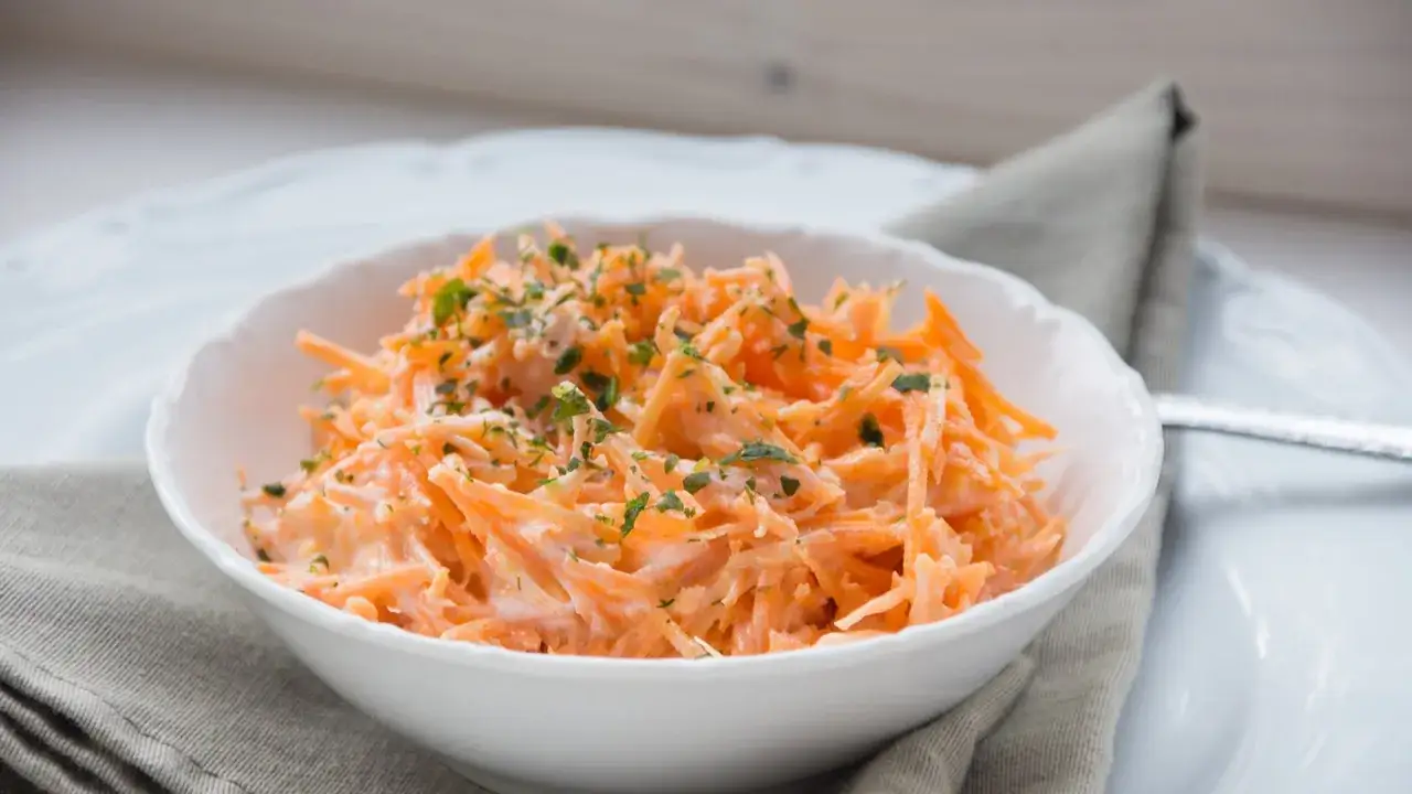 Ein erfrischender Karotten-Apfel-Salat mit Joghurt, garniert mit Petersilie, in einer weißen Schüssel.