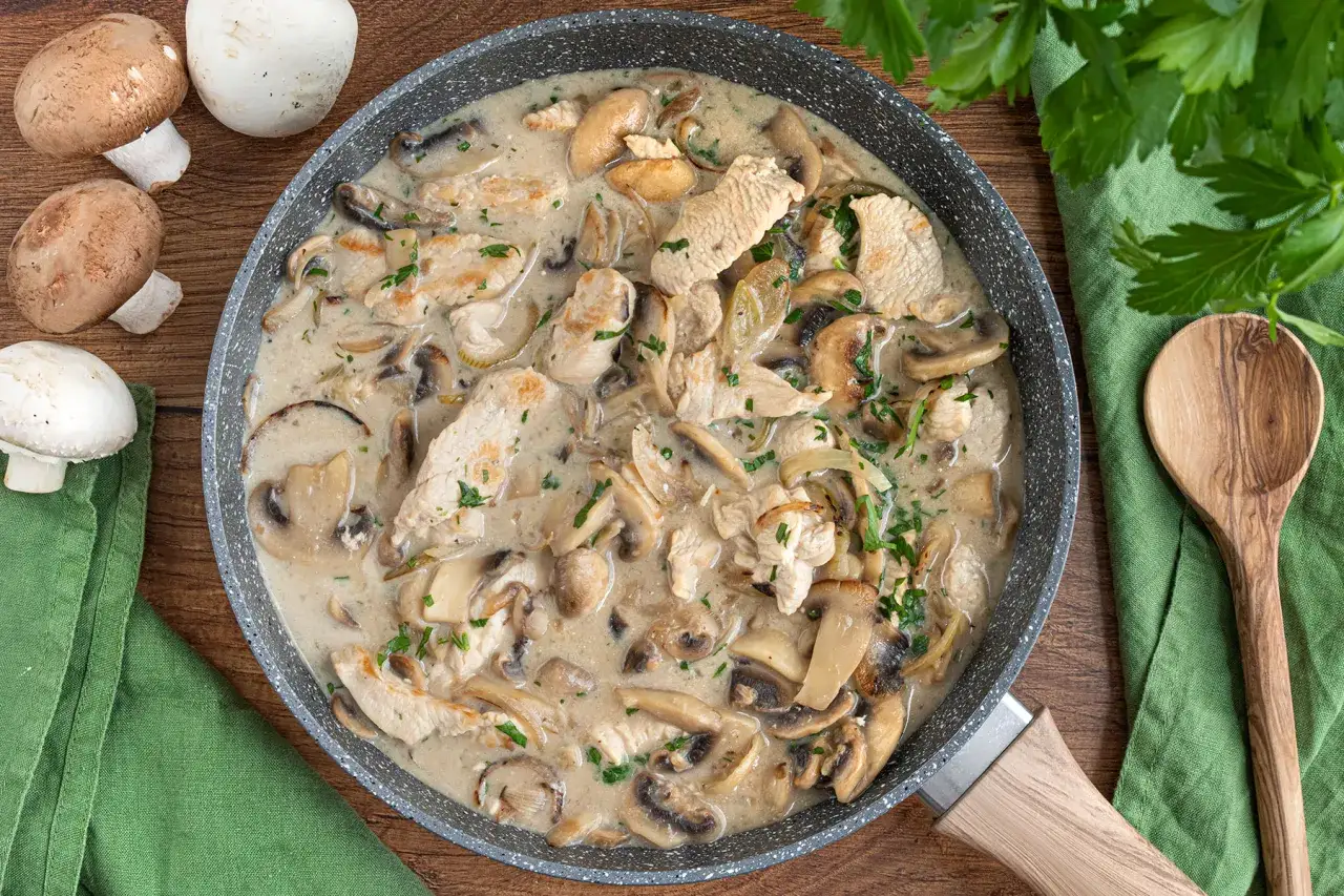 Geschnetzeltes Pute mit Pilzen in cremiger Sauce, garniert mit Petersilie, in einer Pfanne. Daneben frische Pilze und ein Holzl&ouml;ffel.