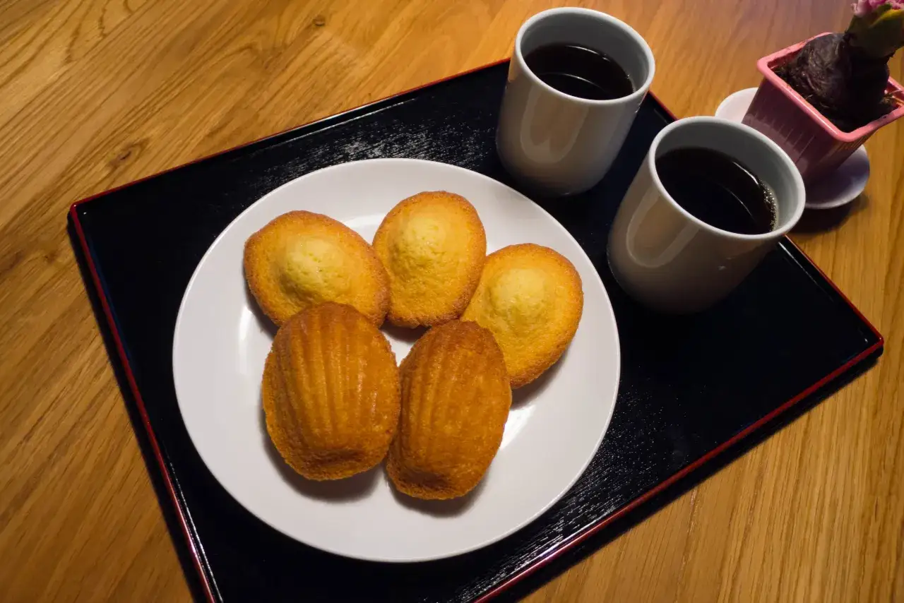 Fünf goldbraune Madeleines auf einem Teller, dazu zwei Tassen Kaffee und eine kleine Pflanze. Perfekt zum Madeleines essen.