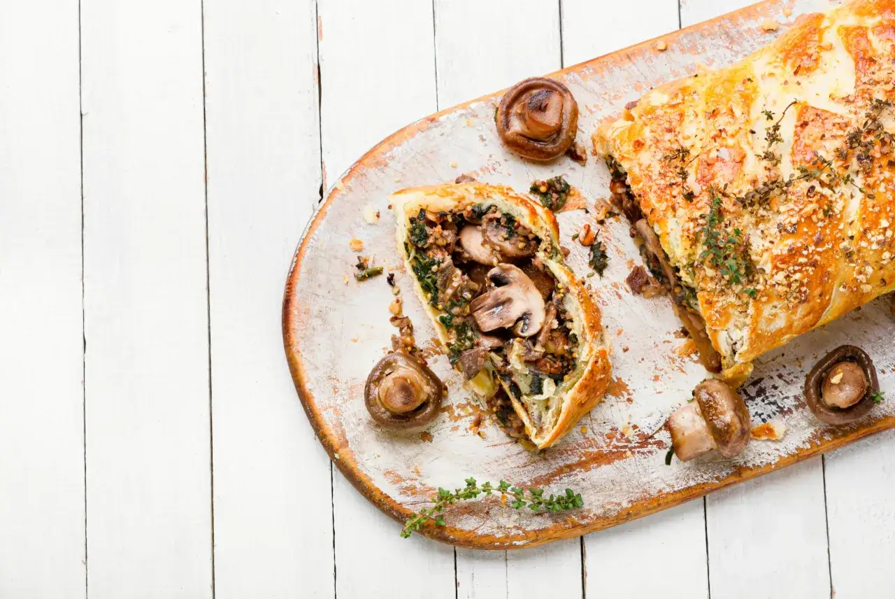 Ein k&ouml;stliches Beef Wellington, aufgeschnitten, mit Pilzen und Kr&auml;utern garniert, auf einem rustikalen Holzbrett.