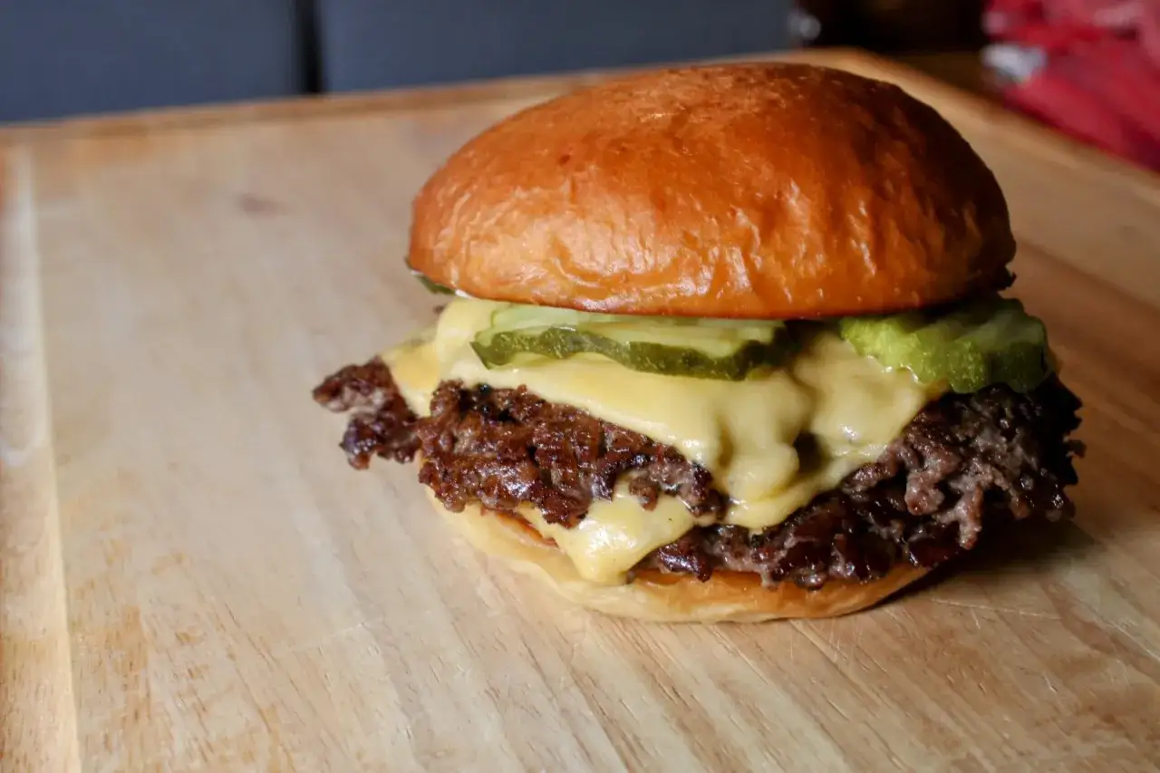 Ein saftiger Smash Burger mit geschmolzenem K&auml;se und Gurkenscheiben auf einem Holzbrett. So sehen leckere Smash Burger aus!