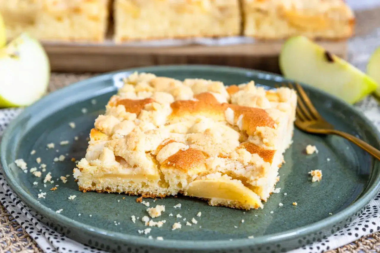 Ein Stück vom besten Apfelkuchen mit Streusel, saftig und goldbraun gebacken, liegt auf einem Teller. Frische Apfelspalten im Hintergrund.
