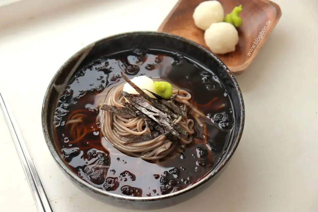 Schüssel mit Mul Naengmyeon, kalter Buchweizennudelsuppe, garniert mit Algen und Wasabi. Daneben kleine Reisbällchen.