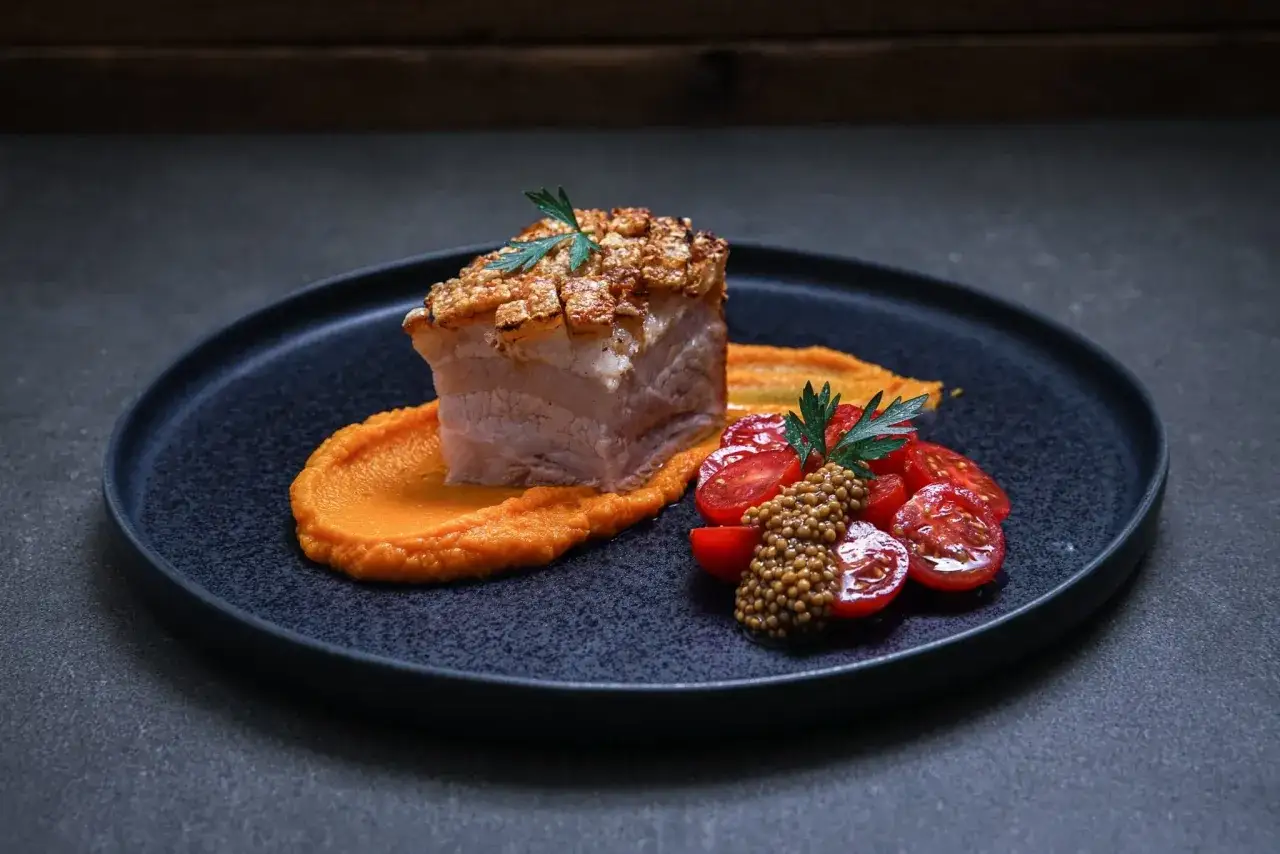 Ein Stück knuspriger asiatischer Schweinebauch auf Kürbispüree, garniert mit Kirschtomaten und Senfkörnern.