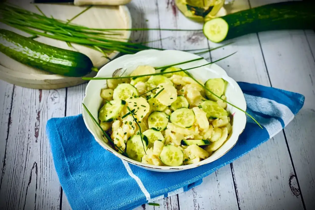 Sch&uuml;ssel mit Kartoffelsalat und Gurkenscheiben, garniert mit Schnittlauch. Frische Gurken und Kr&auml;uter liegen daneben.
