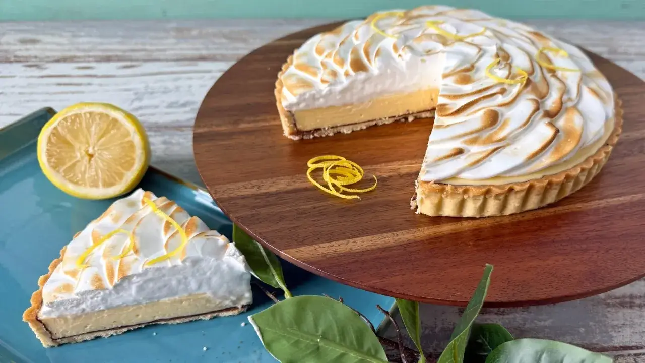 Ein köstliches Zitronentarte Rezept: Ganze Tarte mit Baiserhaube und ein Stück auf blauem Teller, garniert mit Zitronenzesten.