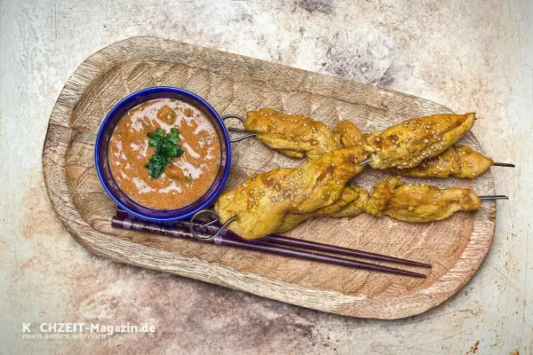 Saftige Satay Chicken Spieße mit Sesam, serviert mit Erdnusssauce und Stäbchen auf einem Holztablett.