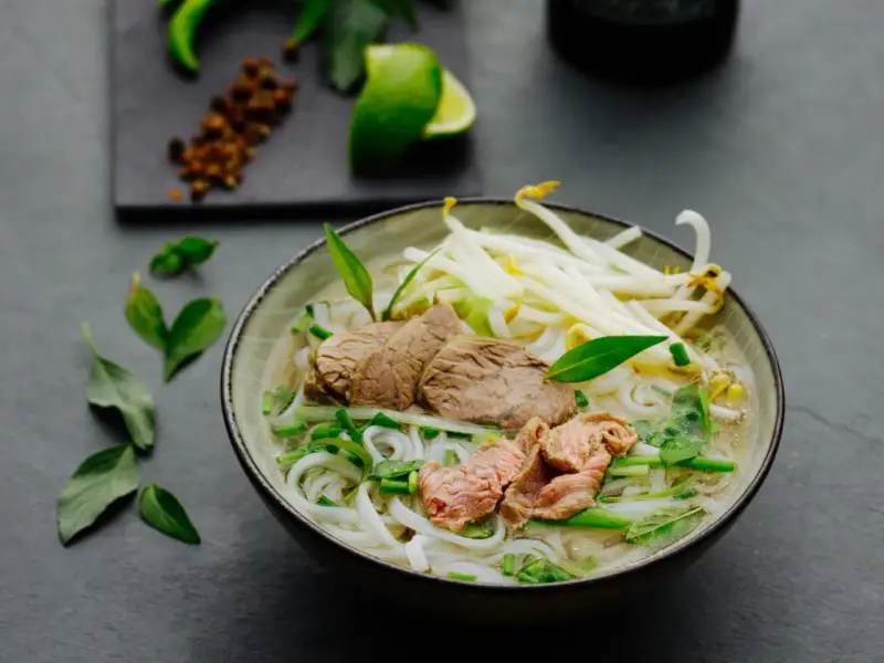 Eine dampfende Schüssel Pho Suppe mit zarten Rindfleischscheiben, Reisnudeln, Sojasprossen und frischen Kräutern. Ein Hauch von Limette und Gewürzen im Hintergrund.